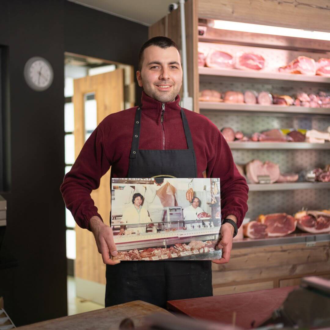 photo of boy holding a picture of his relatives in butcher shop, him standing with refrigerator behind with various meats and cold cuts