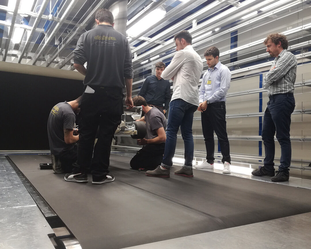 Guys watching dallara employees fitting tires into a skeleton race car