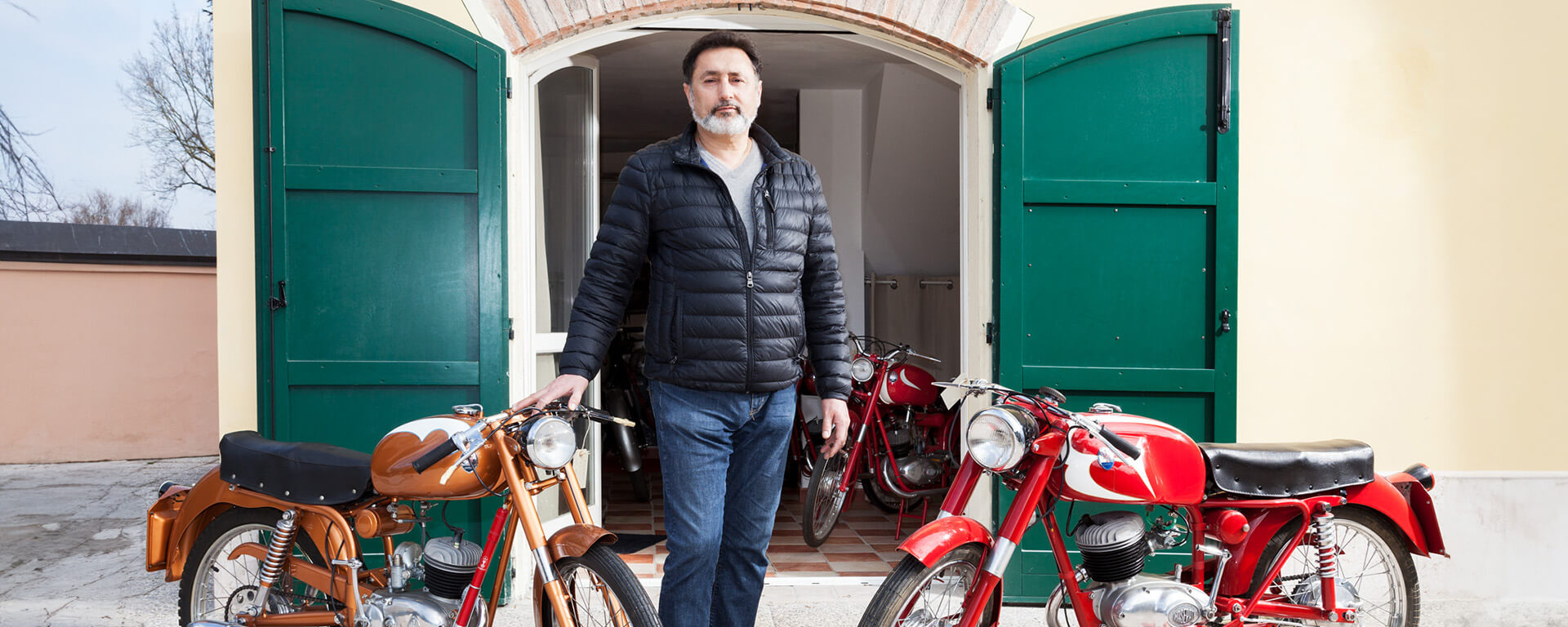 uomo adulto con piumino nero all'esterno di una casa con due moto maserati d'epoca una color bronzo e l'altra rossa