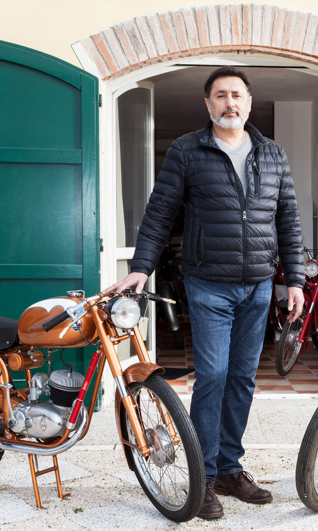 uomo adulto con piumino nero all'esterno di una casa con due moto maserati d'epoca una color bronzo e l'altra rossa