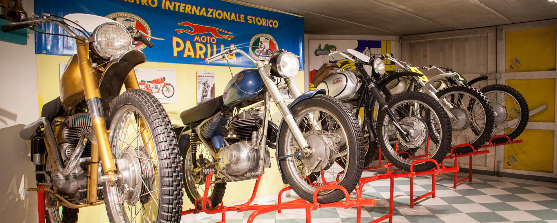 Succession of vintage motorcycles inside an exhibition hall of the Parilla Collection, with the Moto Parilla billboard and logo in the background