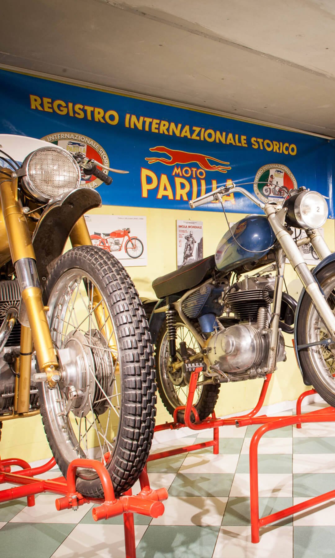 Succession of vintage motorcycles inside an exhibition hall of the Parilla Collection, with the Moto Parilla billboard and logo in the background