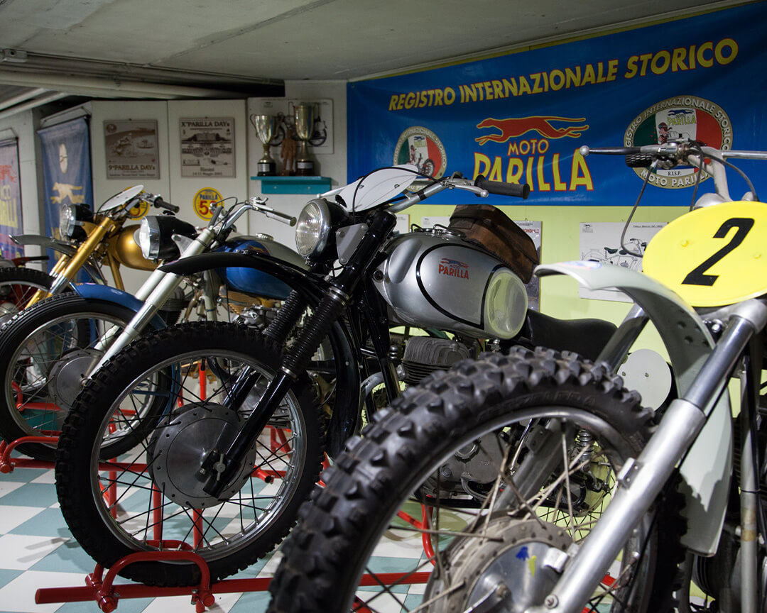 front part of vintage motorcycles in a line-up, inside an exhibition hall of the Parilla Collection, with the Moto Parilla billboard and logo in the background