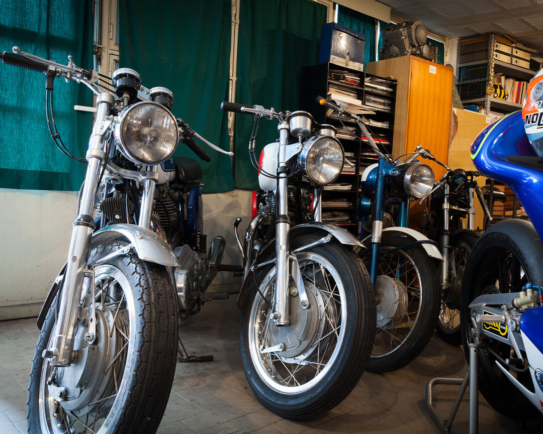 Three vintage motorcycles on display inside the pasquale mesto collection. A blue fairing of another collectible way is glimpsed, with the helmet on the saddle.