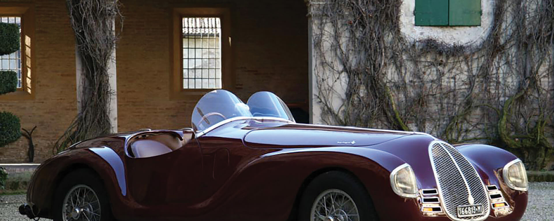 A vintage convertible car of those burgundy, from the righini collection, parked in front of an old building