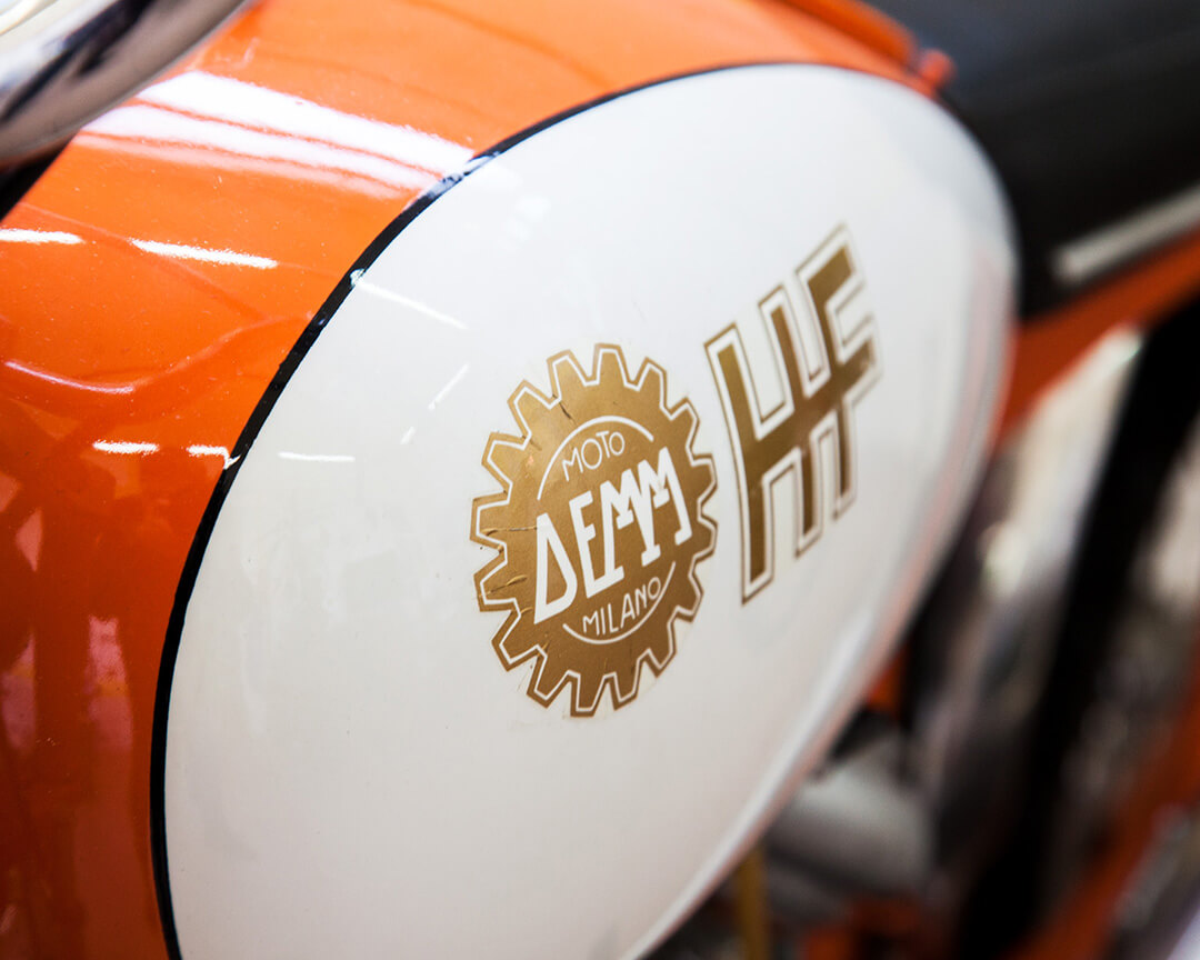 Dettaglio del logo Demm sul serbatoio bianco, arancione e nero di una moto d'epoca del museo Demm