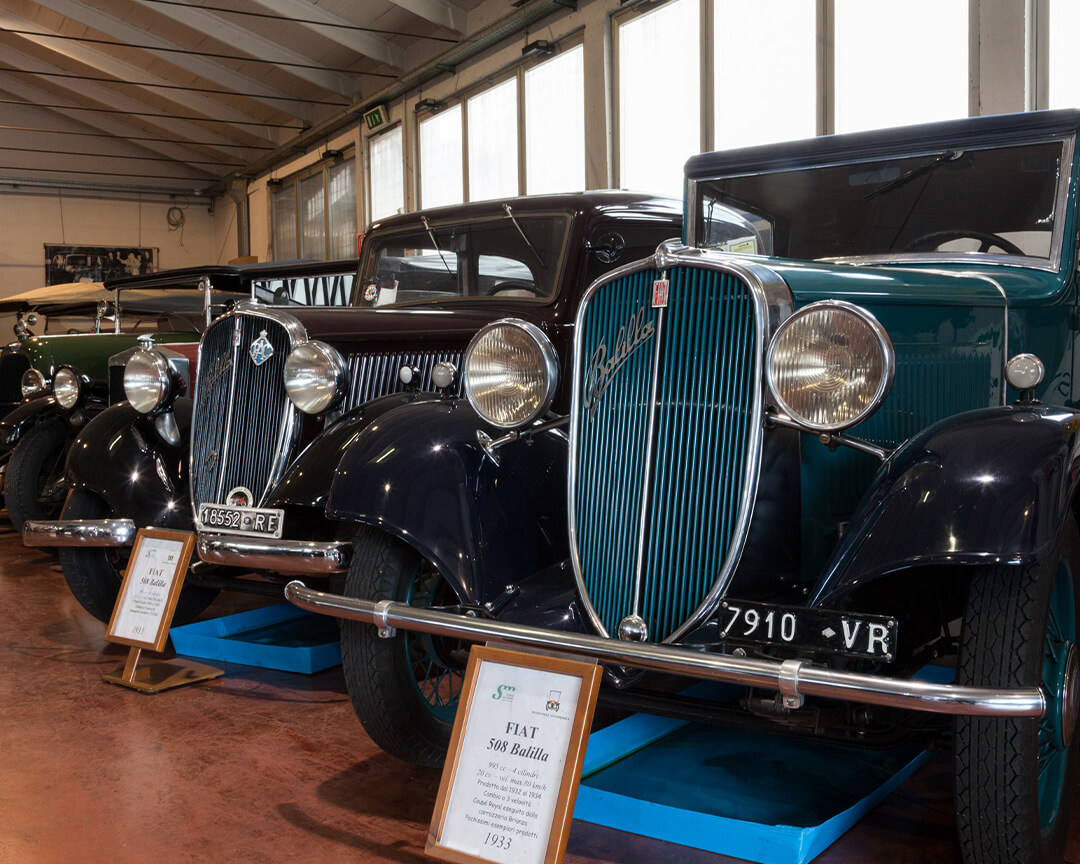 two Fiat 508s from the 1930s parked side by side inside the automobile museum. in the foreground, the headlights and air intake grille are clearly visible