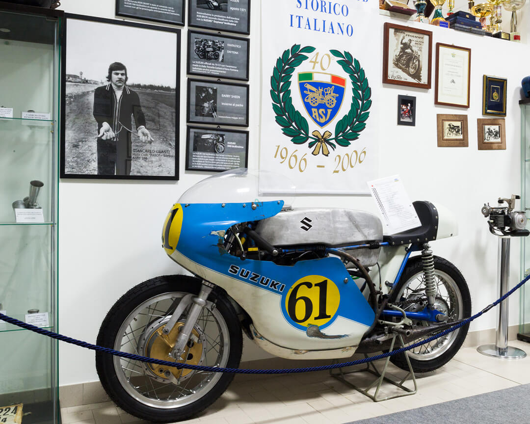 A vintage suzuki track motorbike inside the bariaschi motorbike museum, with photos, plaques and motorbike memorabilia on the wall