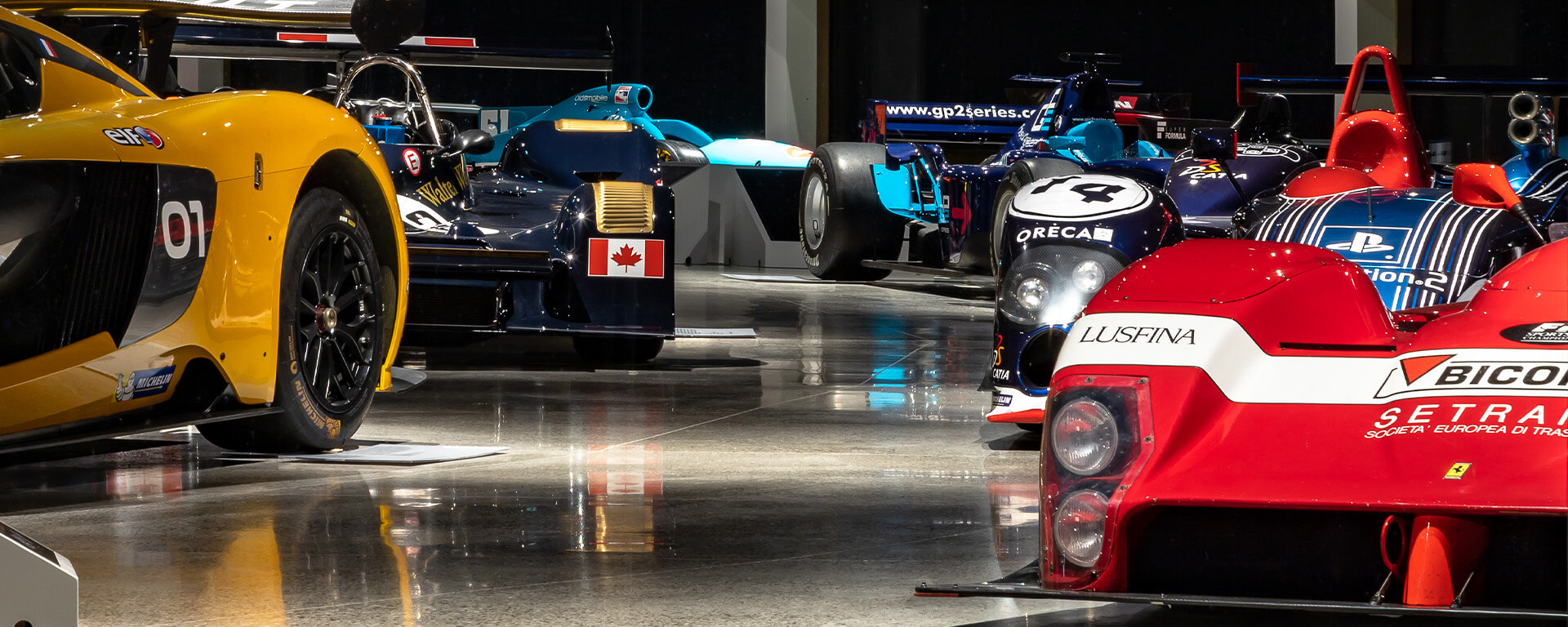 exhibition of racing cars in a museum. In the right foreground is a red sports car with a white fascia, with the inscription ‘Lusfina’ and various sponsor logos, including ‘Bicof’ and ‘Setra’.
