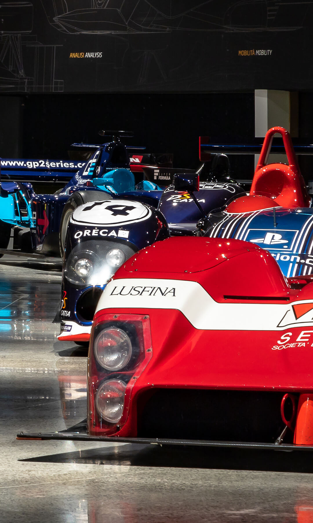 exhibition of racing cars in a museum. In the right foreground is a red sports car with a white fascia, with the inscription ‘Lusfina’ and various sponsor logos, including ‘Bicof’ and ‘Setra’.