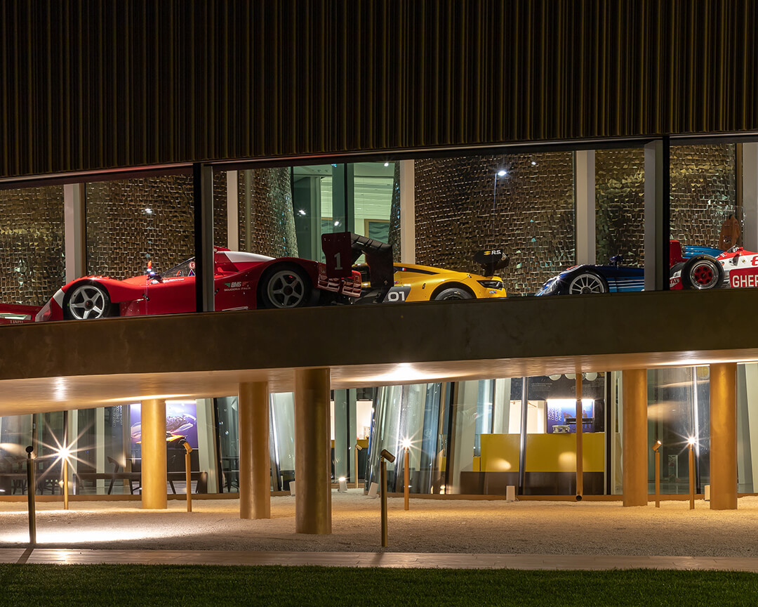 view from the outside at night of the racing cars through the glass of the dallara museum