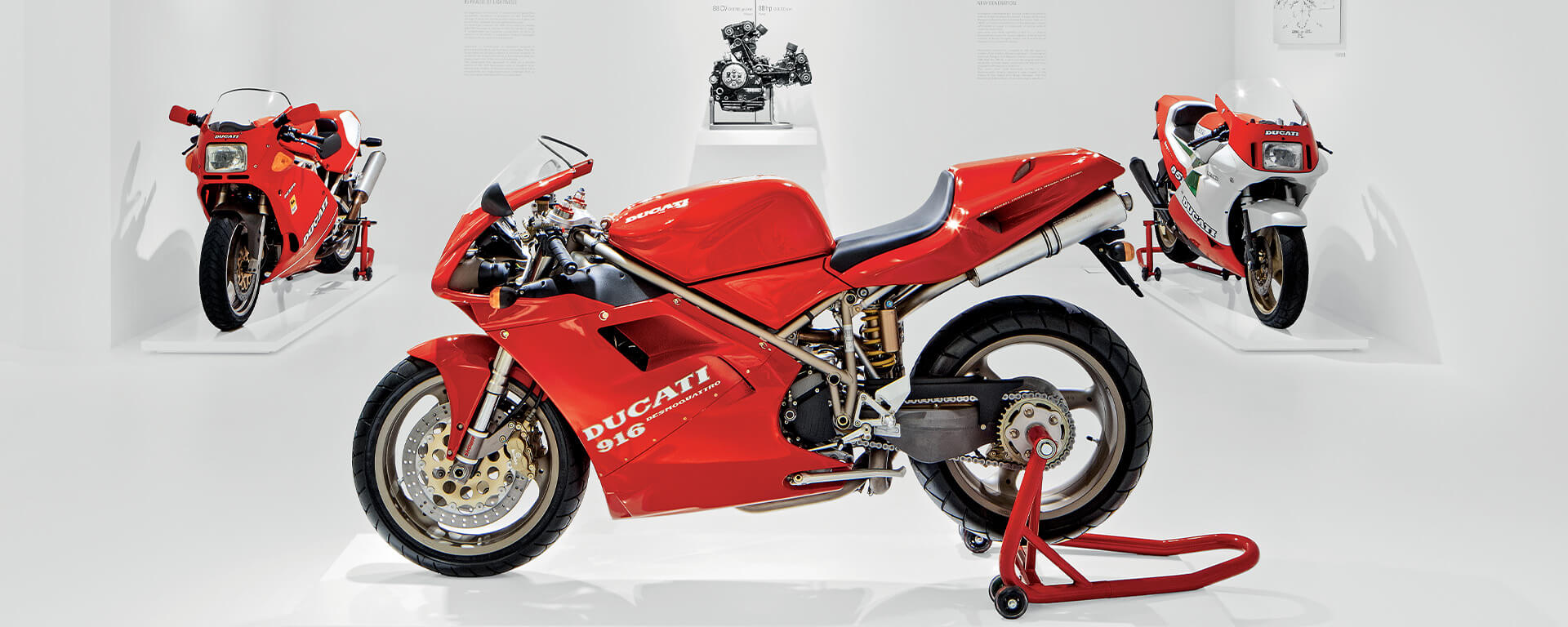 red ducati 916 motorcycle on display , two other motorcycles next to it and a metal motorcycle component in the background