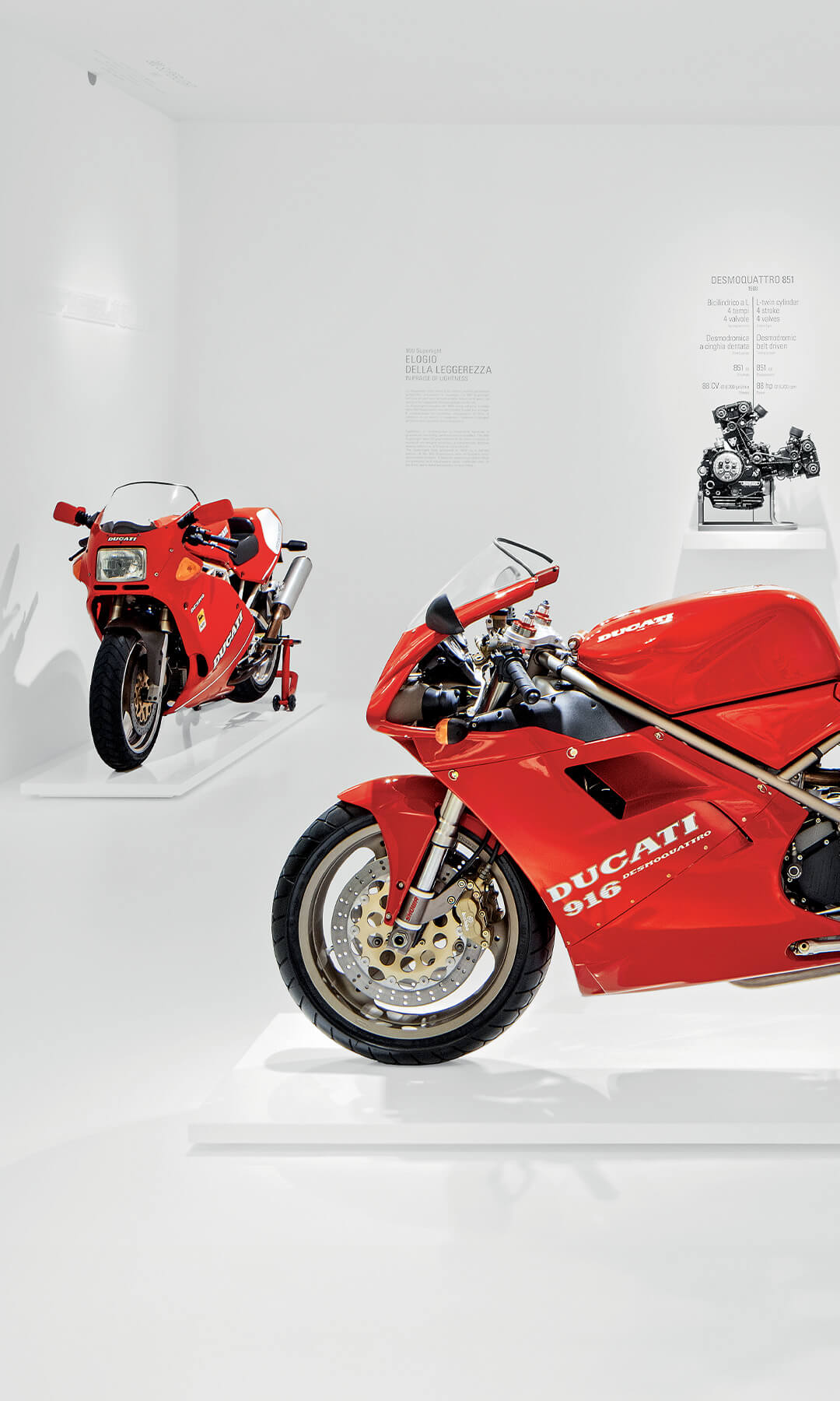 red ducati 916 motorcycle on display , two other motorcycles next to it and a metal motorcycle component in the background