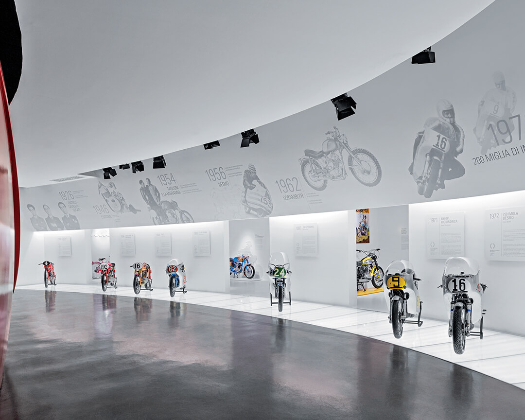 In una sala espositiva della sede ducati sono esposte moto d'epoca che hanno fatto la storia del marchio nel mondo delle corse, la parete bianca ospita date e informazioni riguardante le moto.
