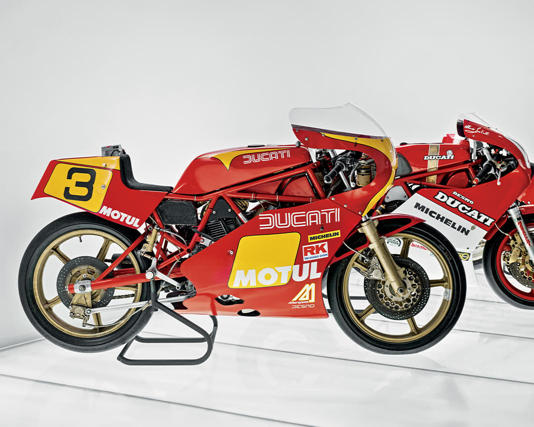 Vista laterale di due ducati da circuito d'epoca. la ducati in primo piano ha una livrea rossa e gialla, con il numero tre, la seconda moto in secondo piano ha invece una livrea bianca e rossa