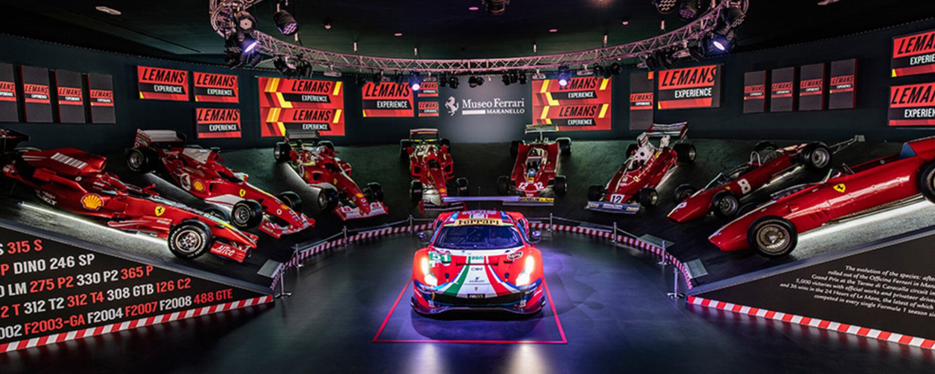 sala del museo Ferrari a Maranello con auto sportiva al centro e attorno diversi veicoli di formula 1 dal più antico al più recente