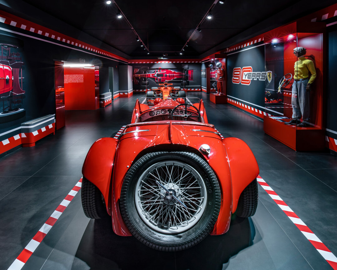 rear view of a vintage ferrari, note the car's large spare tire in the foreground. On the walls ferrari accessories and photos pertaining to the ferrari museum in maranello