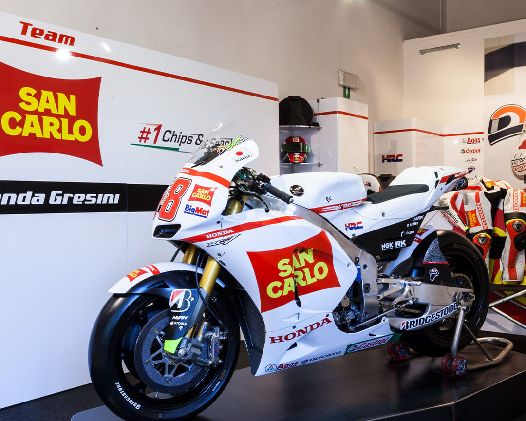 Moto Honda con loghi San Carlo, BigMat numero 58 su piedistallo, dietro pannelli con logo San Carlo e la scritta Honda Gresini. Sullo sfondo caschi e tute da moto