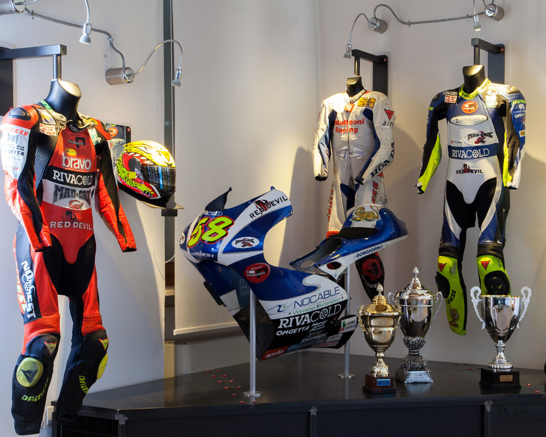 corpo della di una moto con numero 58, accanto coppe in oro e argento e tute da corse di moto