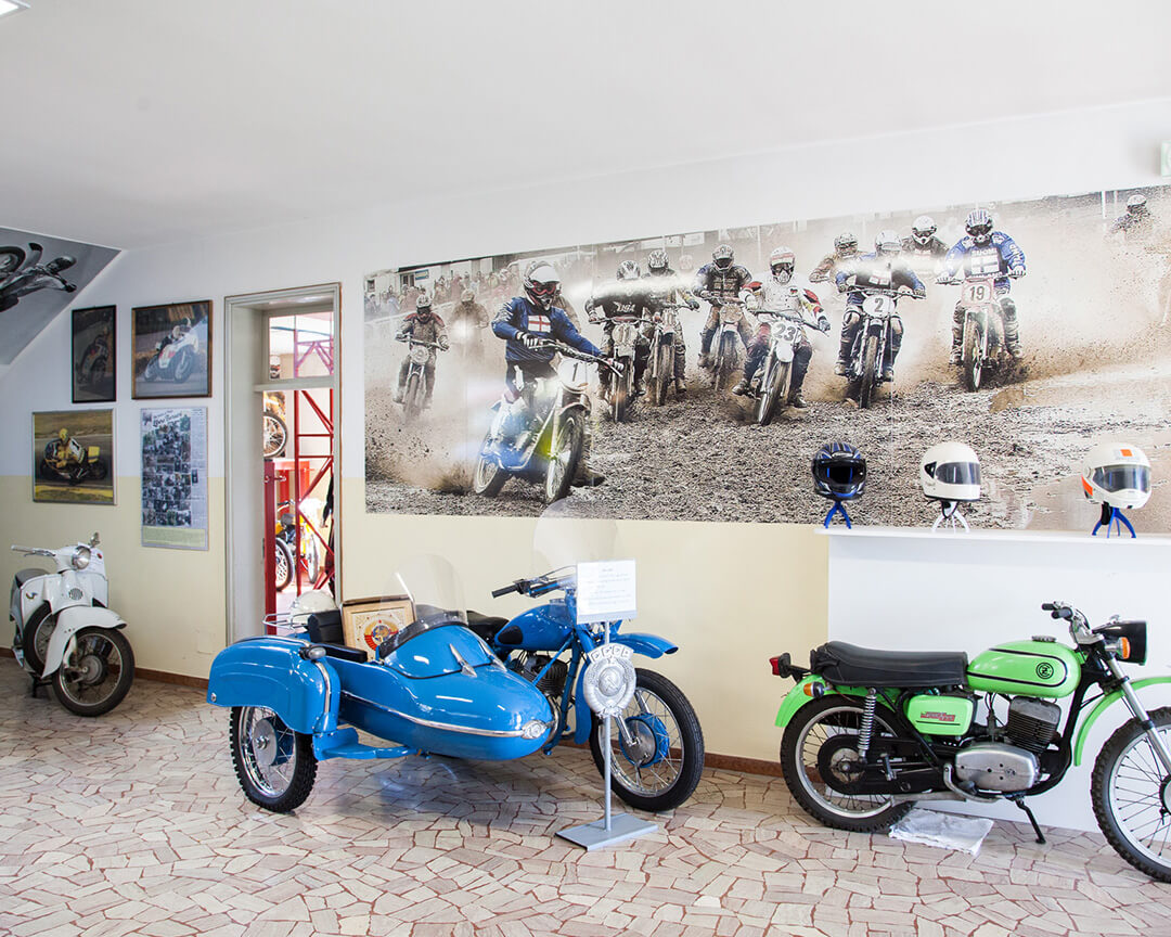 Inside a room at the motorcycle museum in Scortichino, two vintage motorcycles and a collectible blue sidecar are displayed; on the wall is an old photo of a motocross motorcycle race