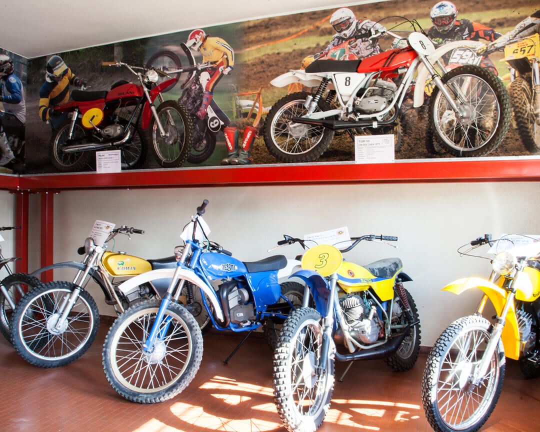 A series of vintage dirt bikes , displayed both above and below a display platform, inside the motorcycle museum in scortichino