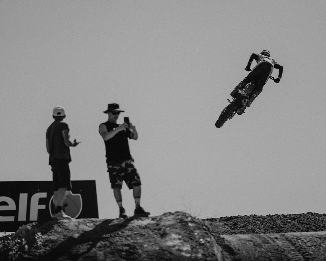 black and white image of motocross rider making a jump and two spectators looking at and taking pictures