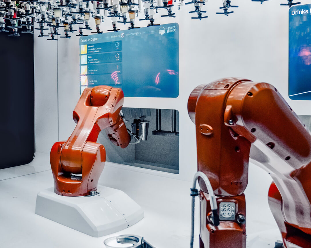 Bar automatizzato con bracci robotici che preparano bevande. Ci sono dosatori per liquidi e uno schermo digitale visualizza gli ordini in coda