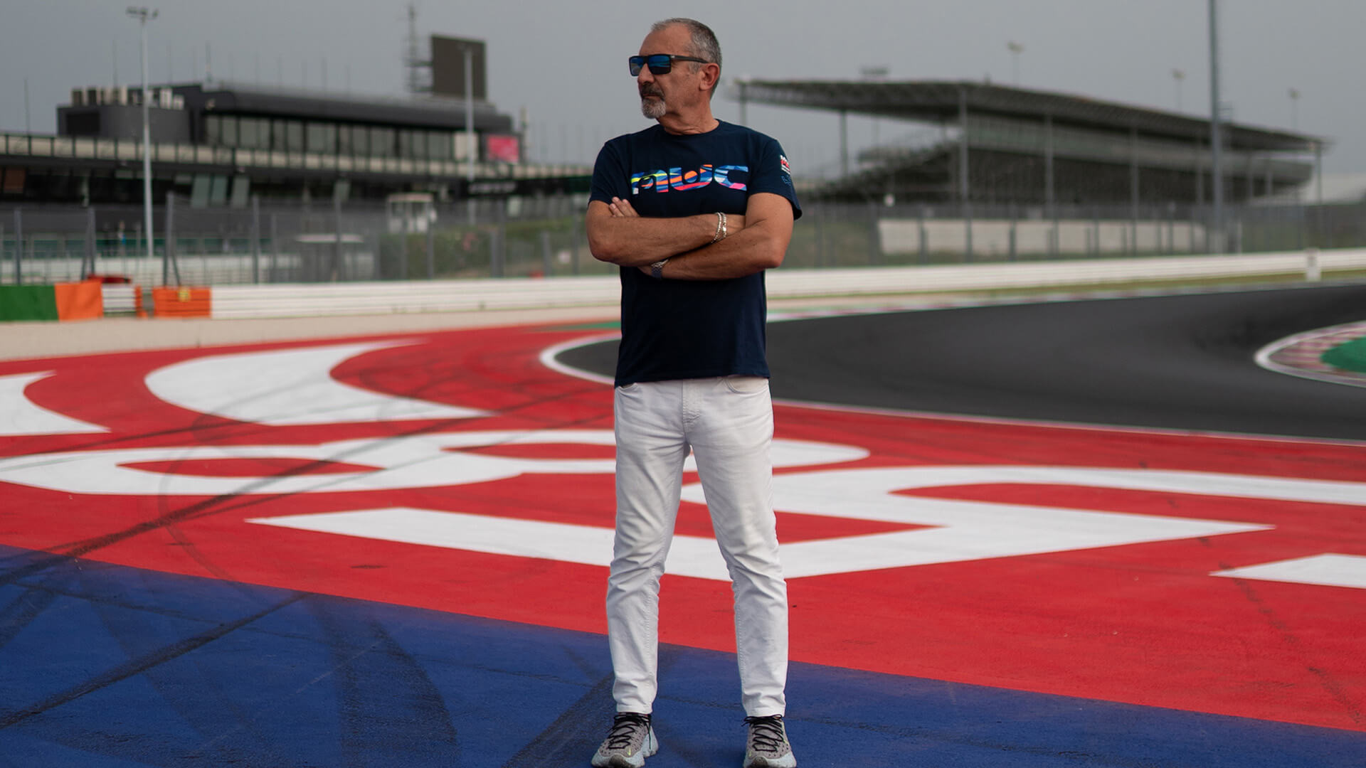 foto di Aldo Drudi in piedi con le braccia incrociate e sullo sfondo il circuito di Misano