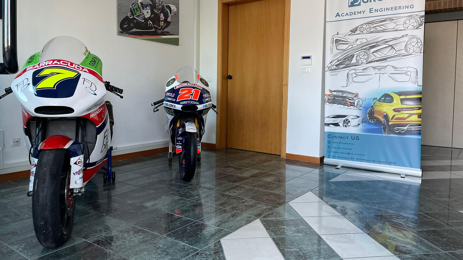 Hall inside FD Group headquarters, with two track motorcycles on display, on the right an FD Group billboard with sketches of sports cars