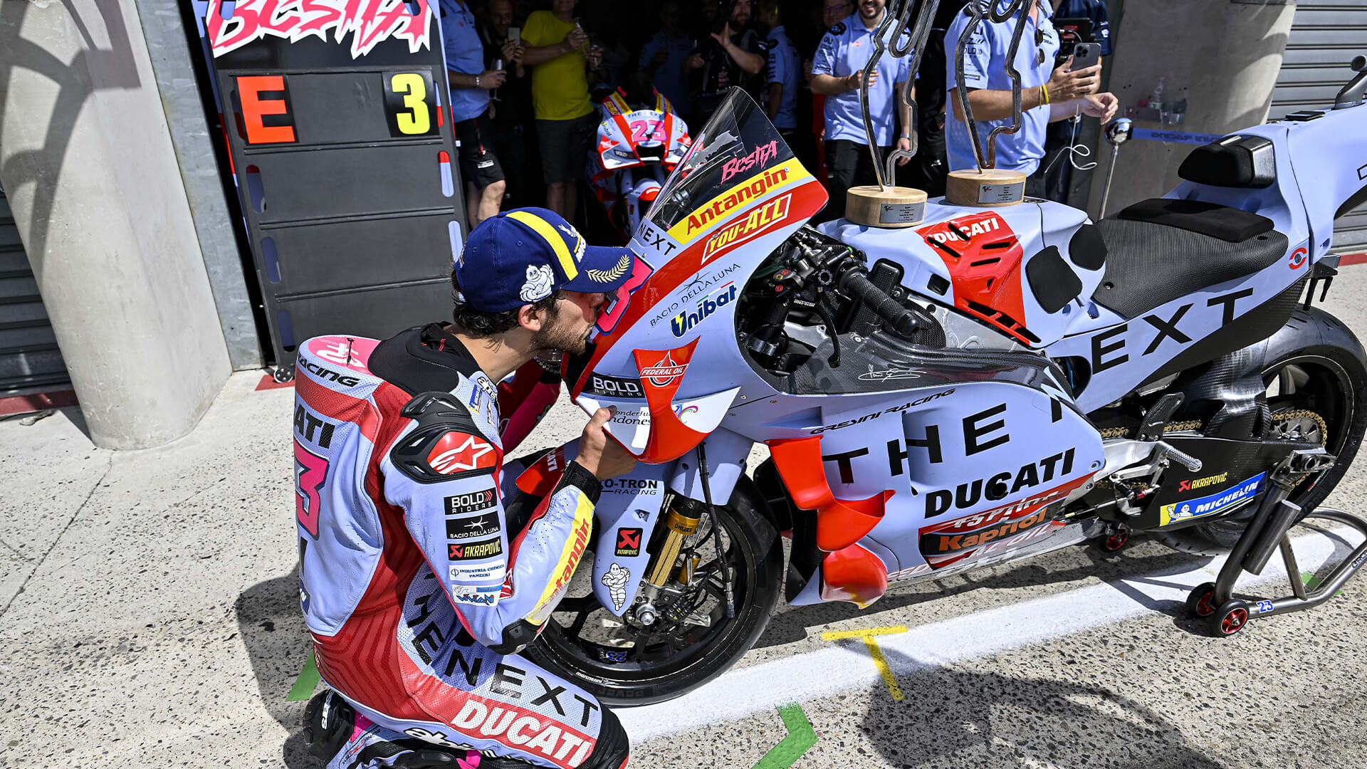 Gresini Racing rider, Enea Bastianini, kneeling in front of his bike, kissing the nose of the vehicle, on top of which are resting two trophies from a MotoGp grand prix