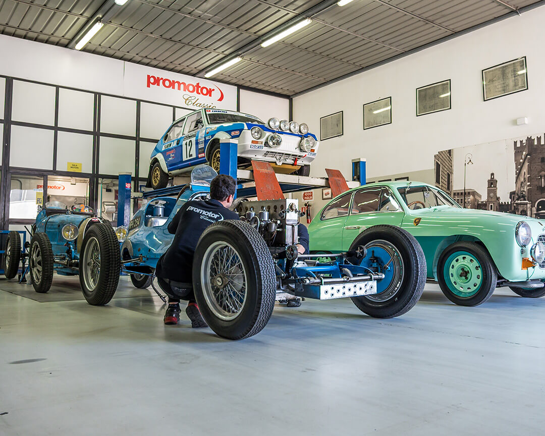 in the foreground a Promotor mechanic working on the engine of, a disassembled classic car, around him three other collector cars, inside the Promotor workshop