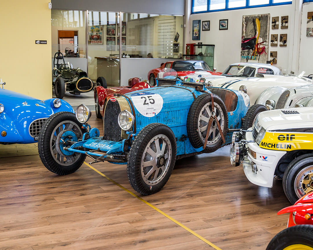 Inside a Gruppo Promotor exhibition hall, a Bugatti Type 35, the nose of a Renault 5 turbo and the front end of a blue Stanguellini can be seen in the foreground.