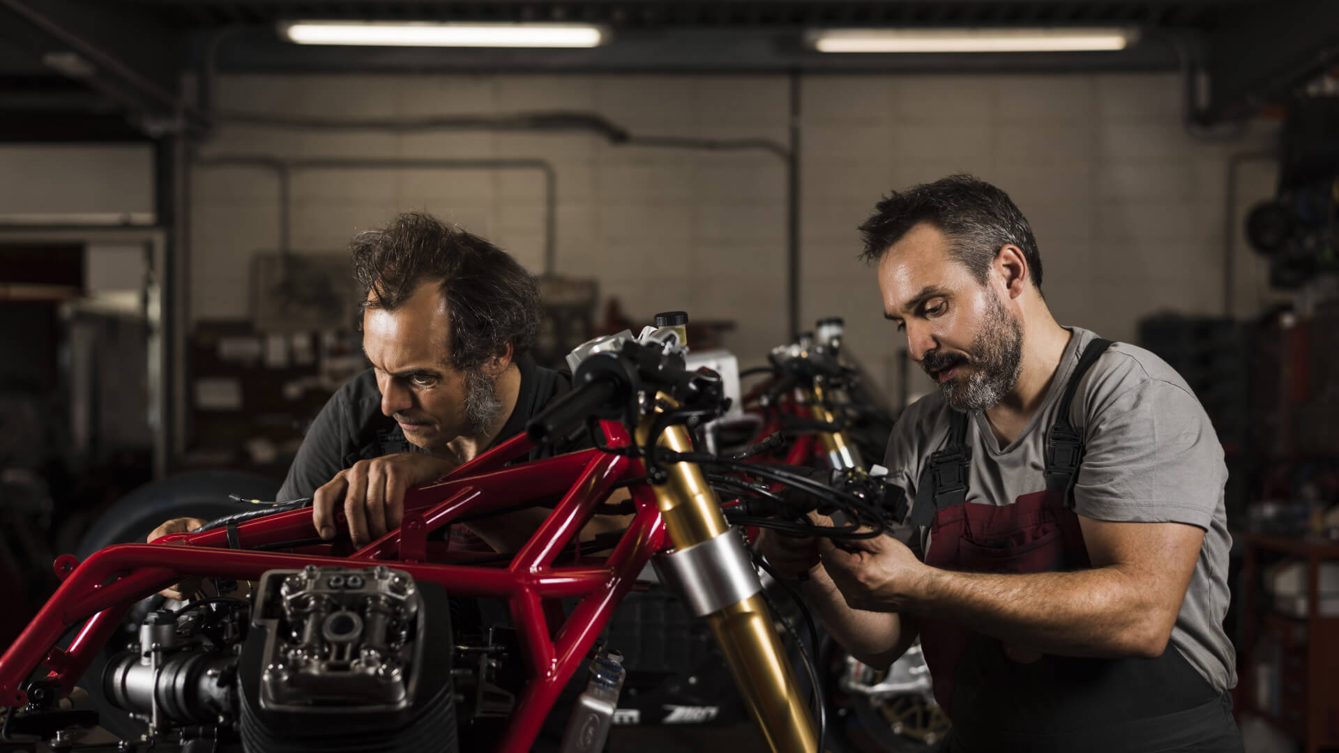 Two mechanics work on a motorcycle without a fairing with the frame exposed, inside the Guareschi workshop
