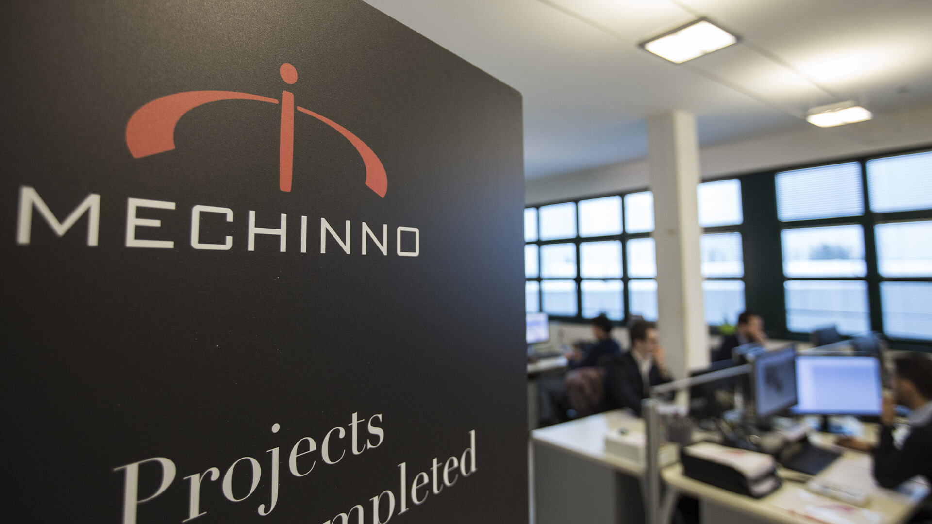 Mechinno logo in the foreground and in the blurry background employees working at their desks