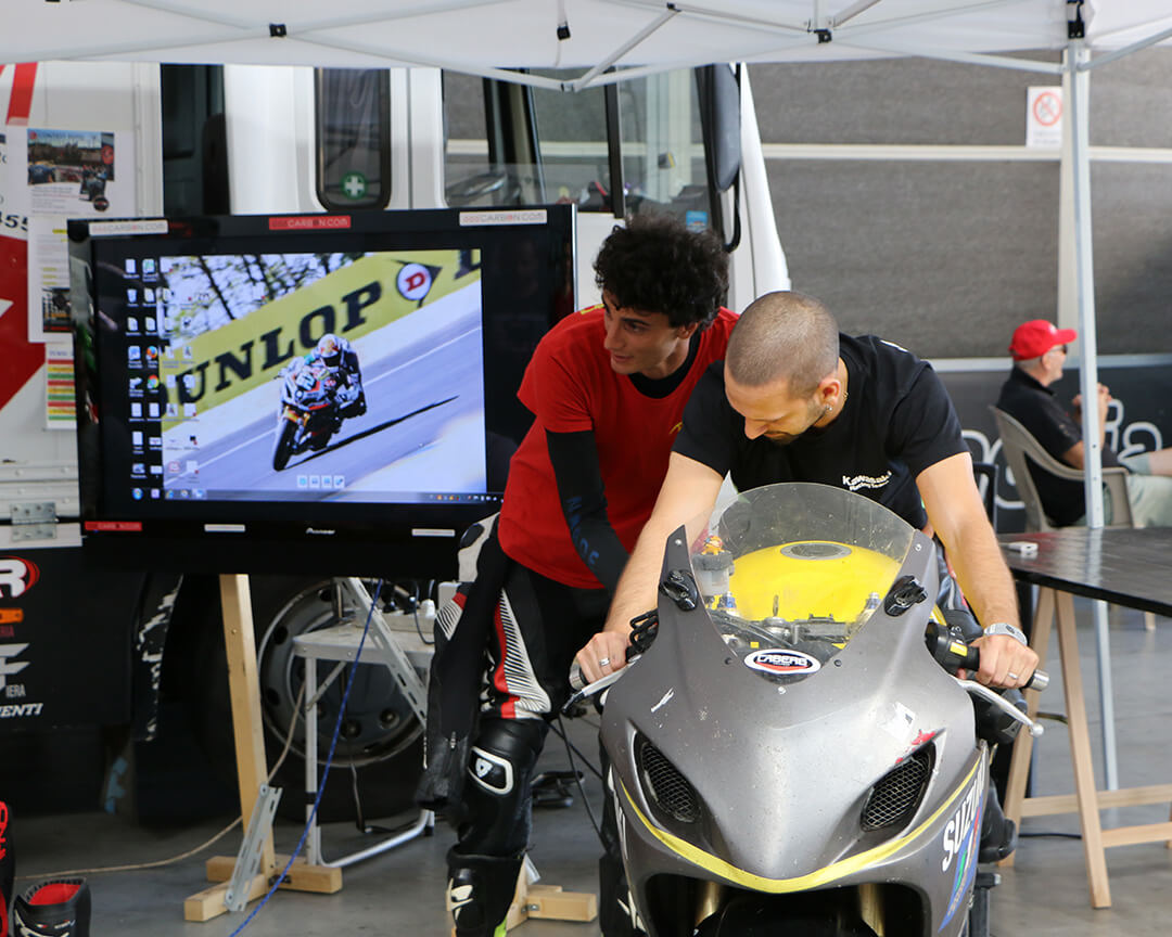 On the screen a motorcycle rider and two boys one explaining and the other on top of the motorcycle