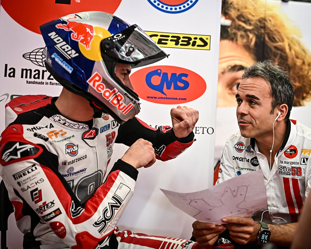 pilota con casco con logo Redbull che scherza con il suo allenatore che ha in mano la mappa del circuito, le cuffie con il filo bianco e una polo con il logo SIC 58