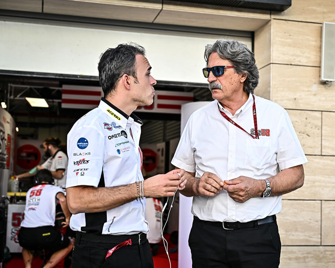 padre di Marco Simoncelli con baffi e occhiali e con maglietta bianca con logo Sic 58 che parla con un collega con tshirt bianca che tiene in mano cuffiette con filo bianco
