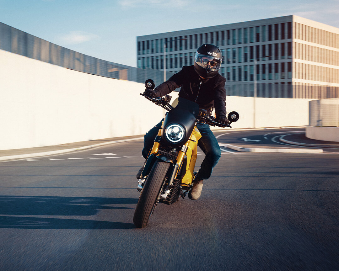 Tazzari electric motorcycle, with yellow paint, being driven by a test rider entering a traffic circle