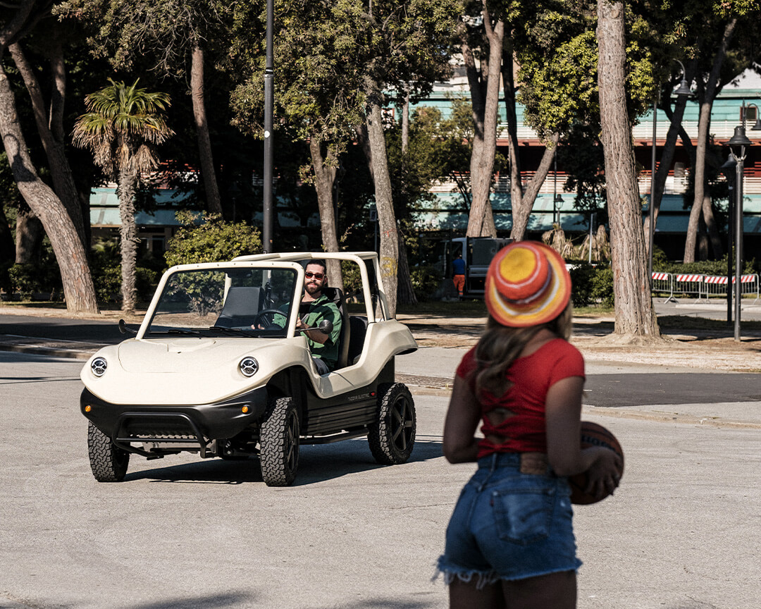 Cream-colored dune buggy tazzari, driven by a boy with glasses, catching up with a girl in a hat