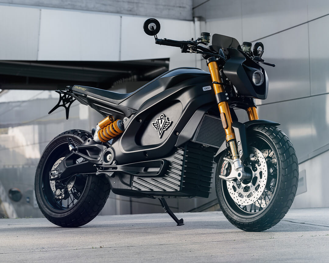 A tazzari electric motorcycle, black in color, parked and leaning on its stand, with a gray building behind it