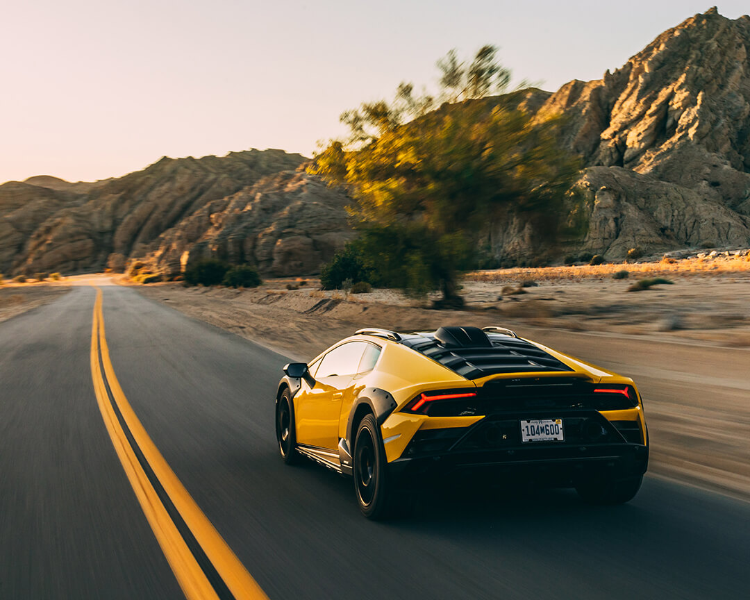supercar Lamborghini gialla che sfreccia lungo una strada deserta circondata da un paesaggio roccioso al tramonto.