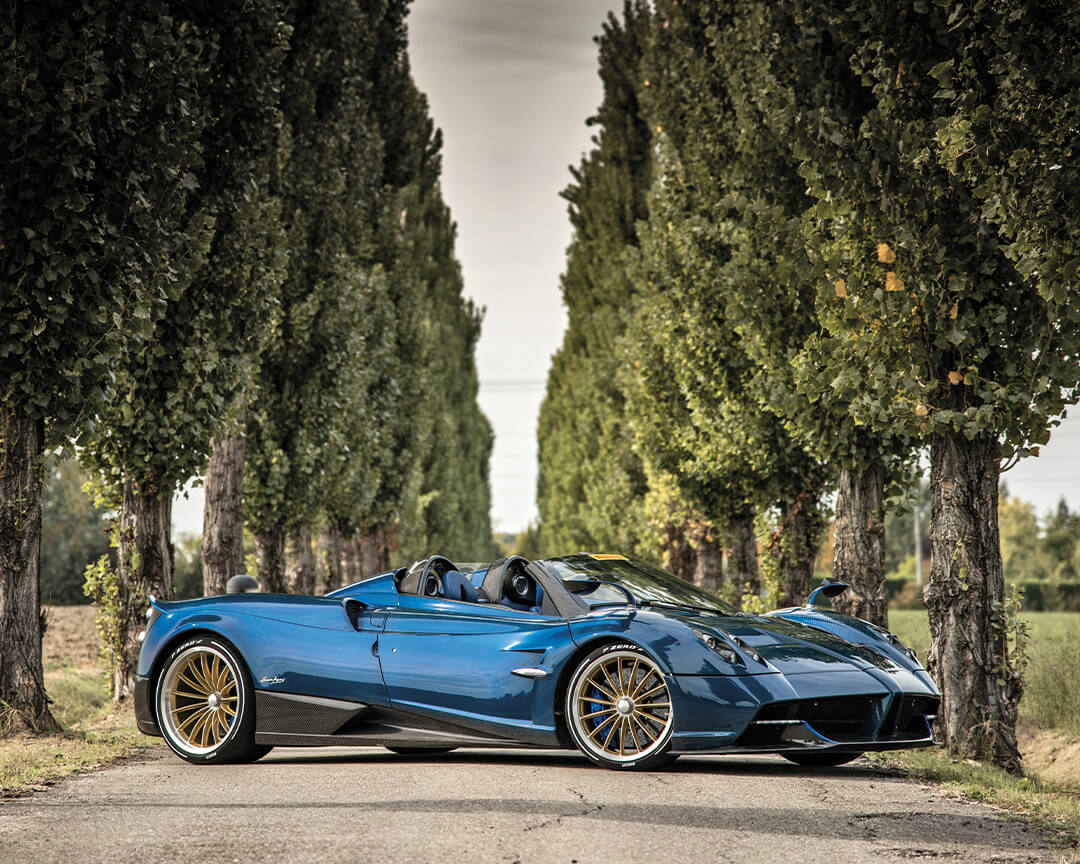 Pagani roadster blu fotografata lateralmente in una via percorsa da cipressi