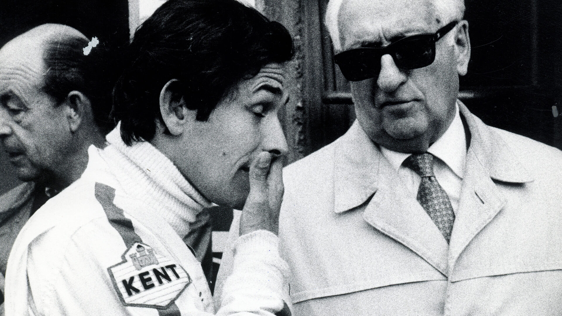 una foto in bianco e nero ritrae un pilota parlare con la mano davanti alla bocca a enzo ferrari
