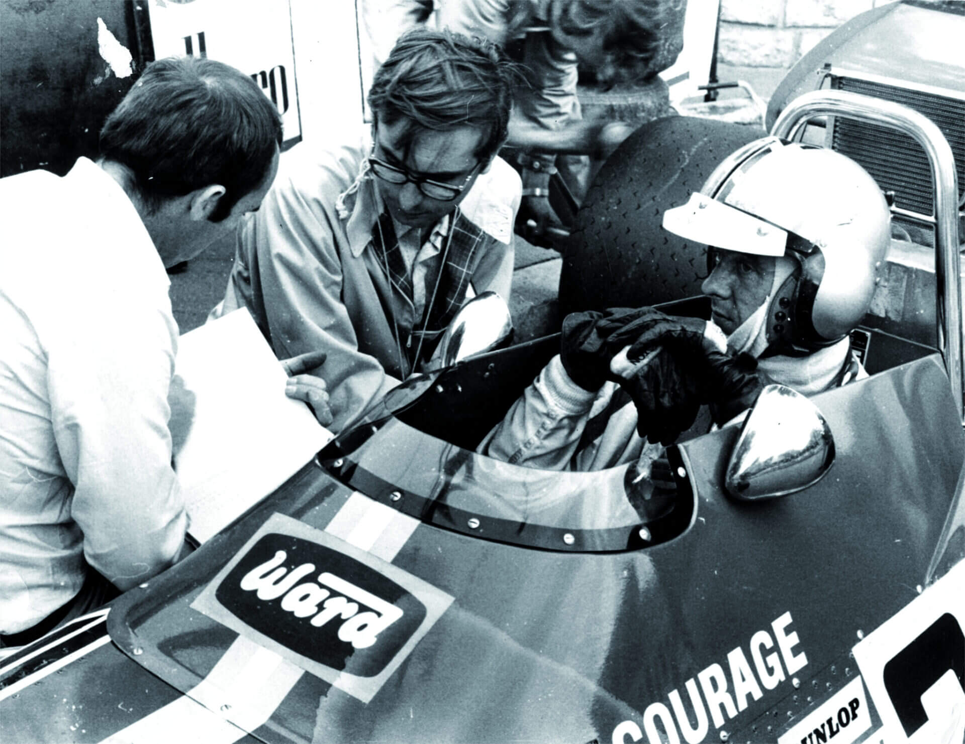 black and white photo of a racing driver in a single-seater car, wearing a helmet and gloves, talking to two team members