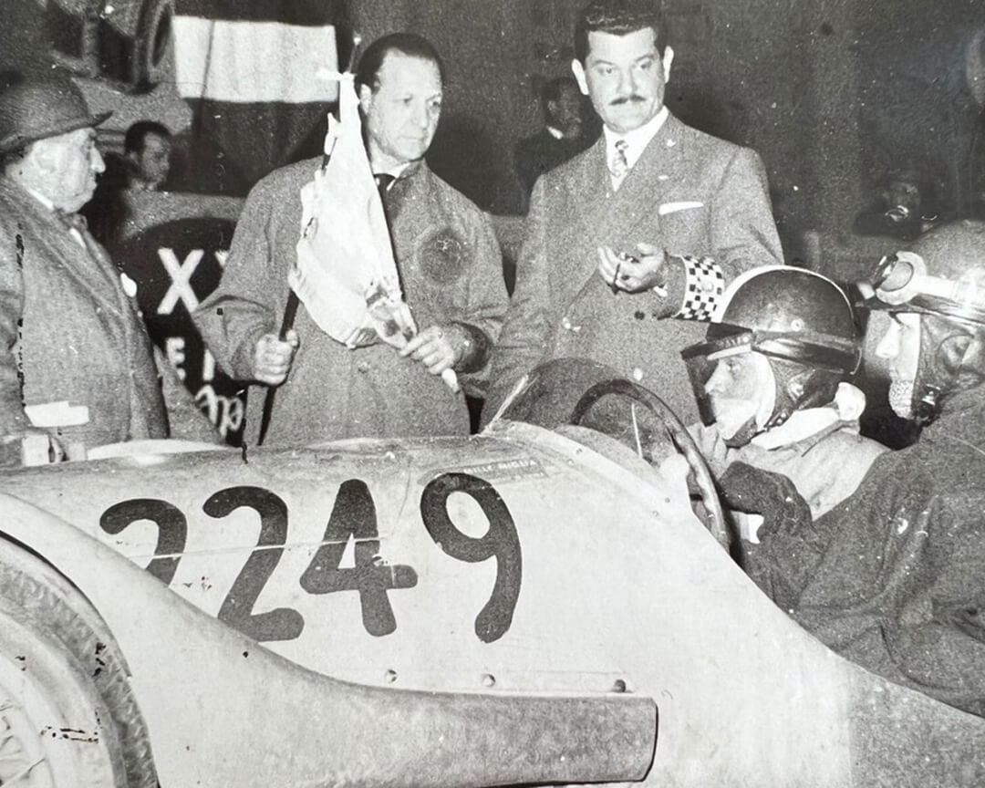 foro in bianco e nero di Ilario Bandini con il meccanico Sintoni alla Mille Miglia del 1953 alla partenza della gara con i giudici con la bandiera del via in mano