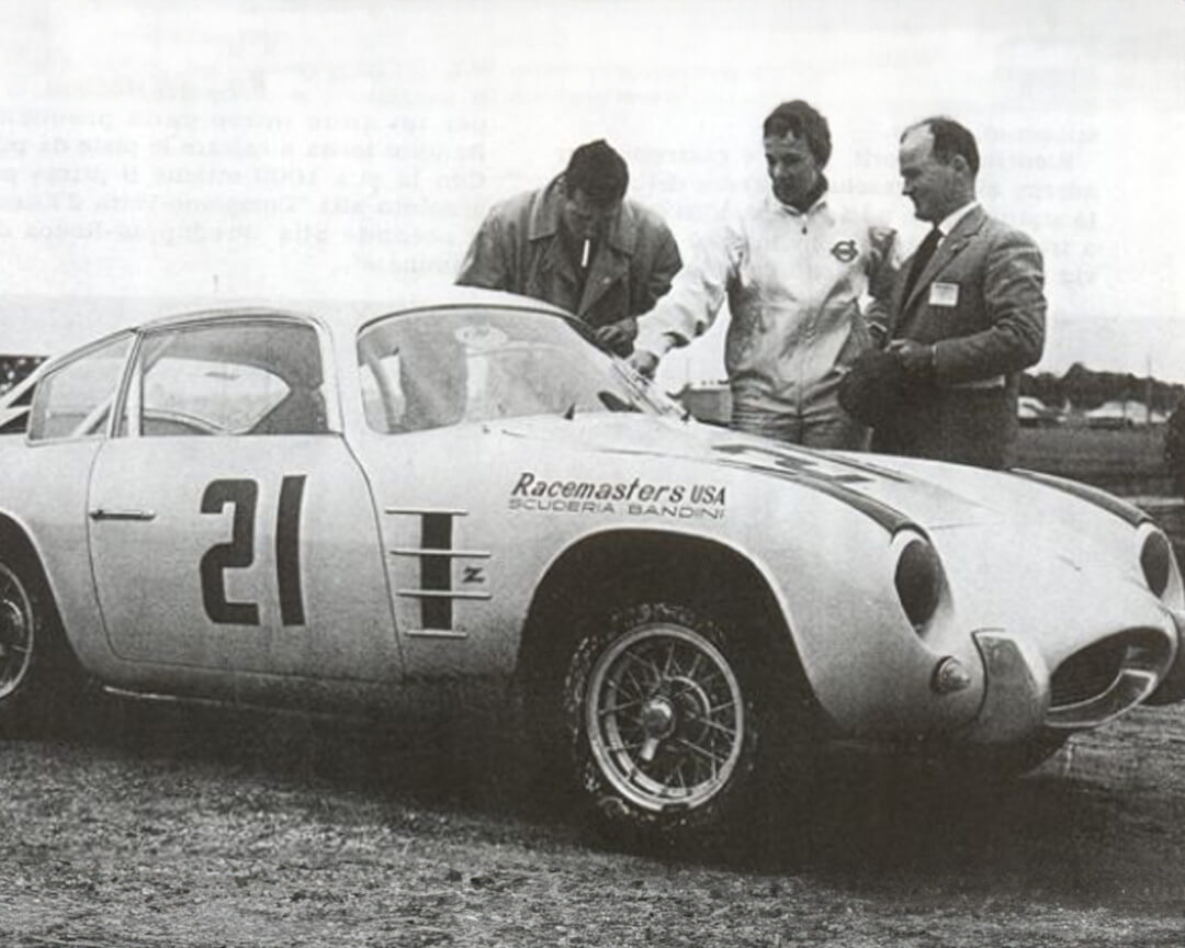 foto in bianco e nero di Ilario Bandini con la sua 750 GT Scuderia Bandini in piedi con il pilota e un altro osservatore