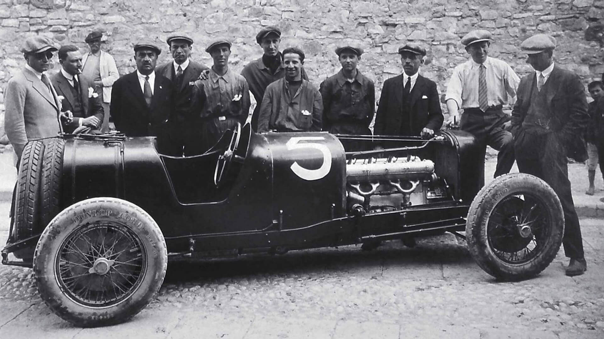 Foto d'epoca in bianco e nero che raffigura una vecchia maserati senza cofano, con dietro meccanici e componenti della famiglia maserati