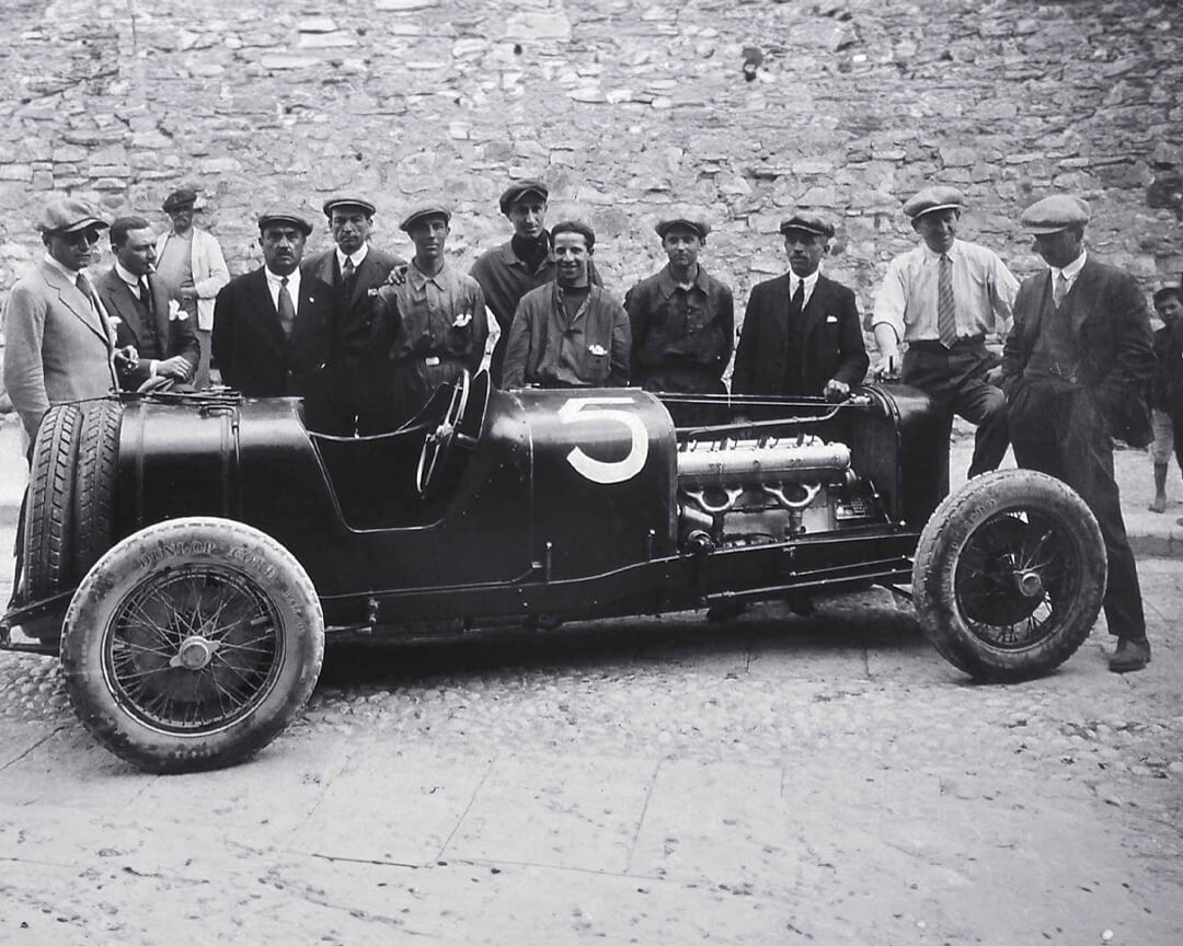 Foto d'epoca in bianco e nero che raffigura una vecchia maserati senza cofano, con dietro meccanici e componenti della famiglia maserati