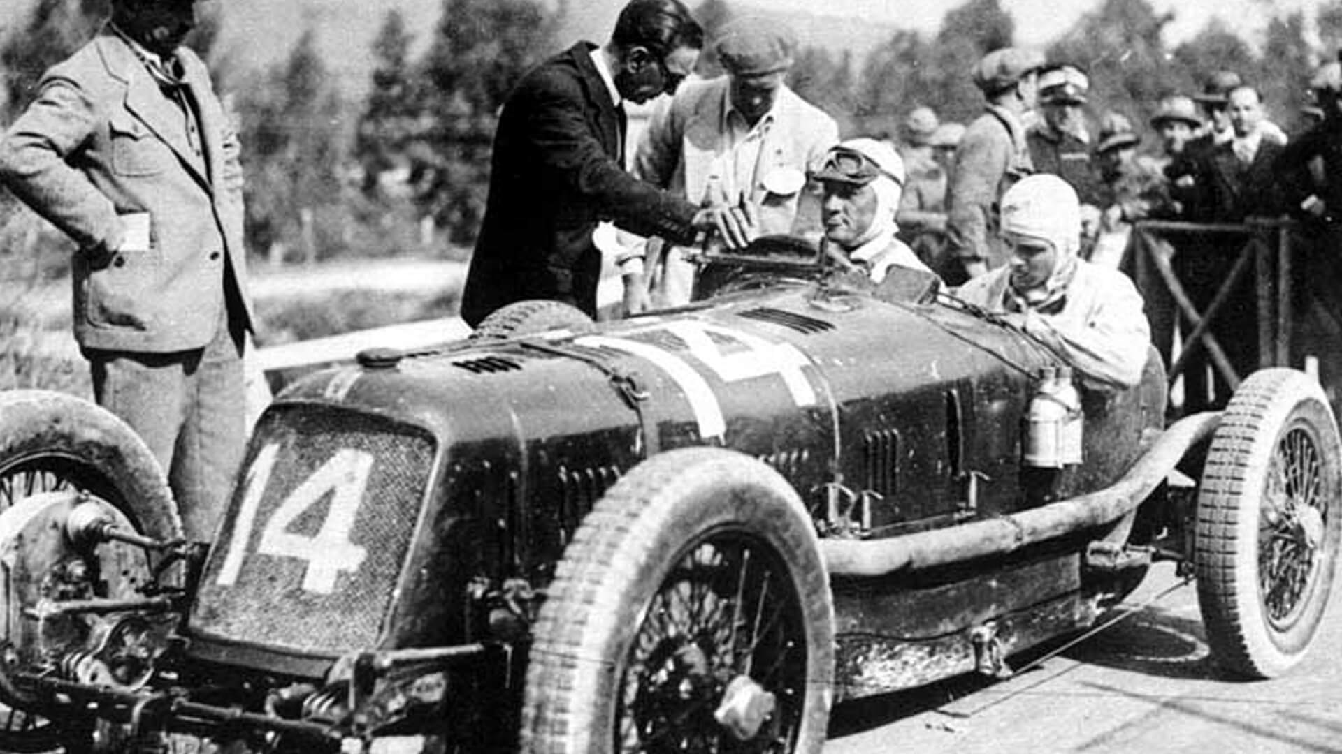 Foto d'epoca in bianco e nero di una vecchia maserati da corsa con il numero 14 e i due piloti alla guida pronti a partire
