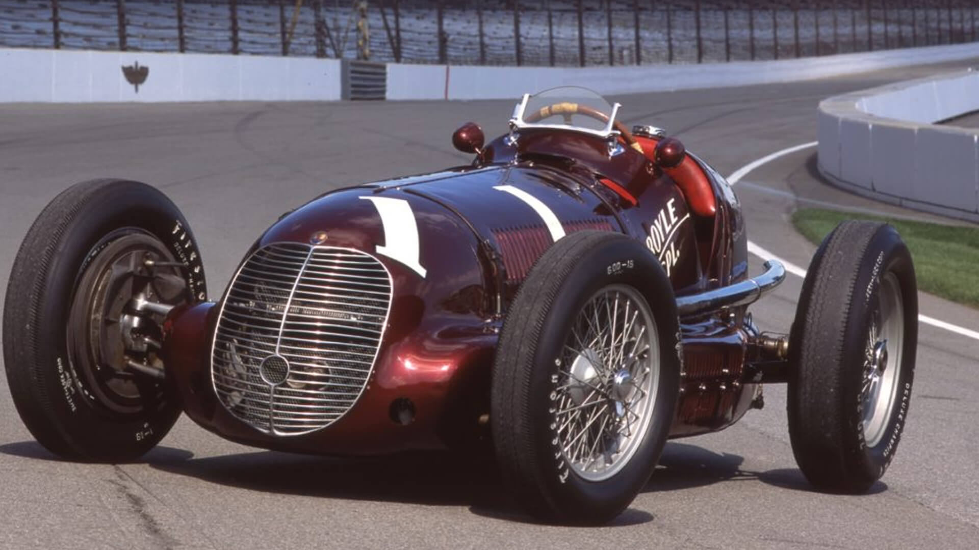 Foto di una maserati d'epoca da corsa, di colore bordeaux e con il numero 1. La vettura è parcheggiata in mezzo ad un tracciato